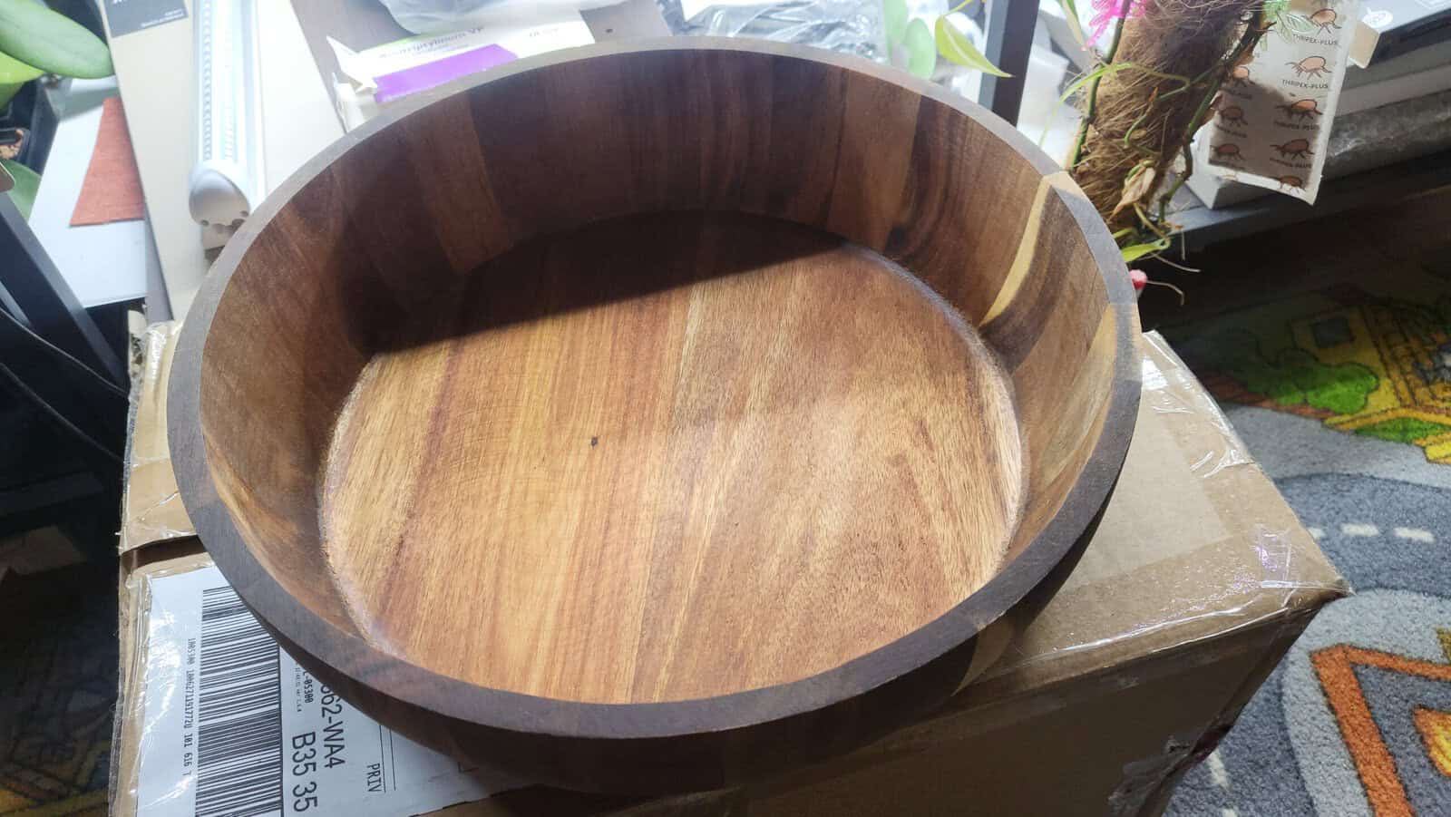 A large, round wooden bowl with a smooth, polished finish, sitting on a cardboard box near a window. The warm, natural grain of the wood is visible, and there are some plants and books in the background.