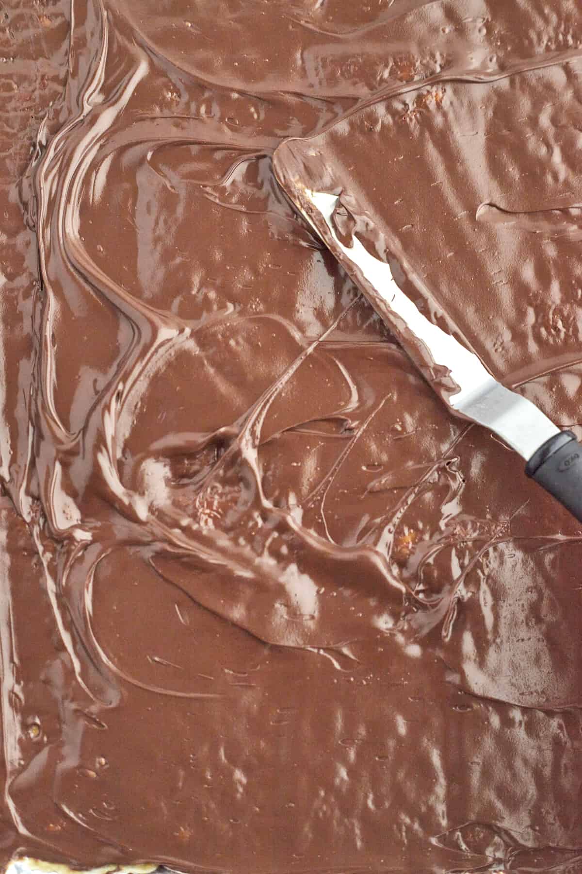 melted chocolate being smoothed over caramel covered saltine crackers to make crack candy