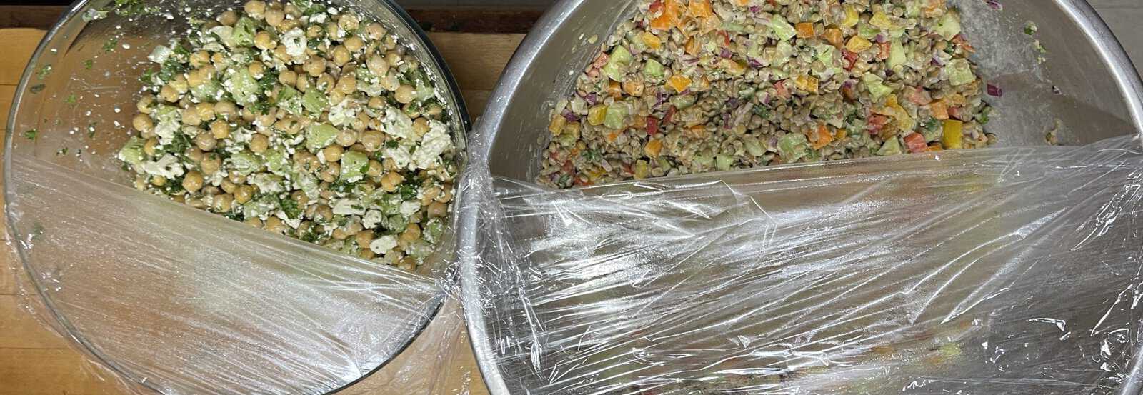 Two bowls of salad covered with plastic wrap. The left bowl contains a chickpea and feta salad with herbs. The right bowl contains a mixed vegetable salad with peppers, onions, and cucumbers. Both are set on a wooden surface.