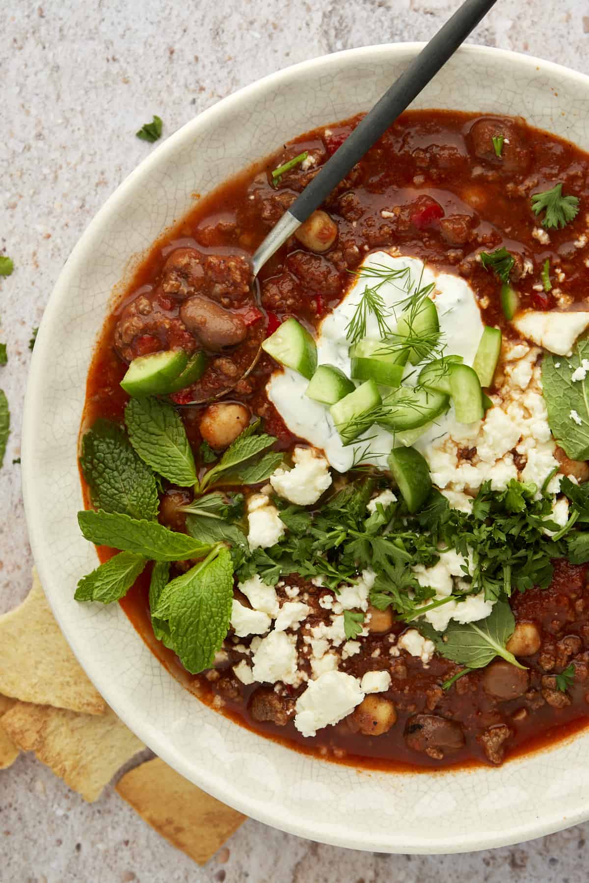 a white bowl full of beef chili topped with yogurt sauce, feta, and herbs