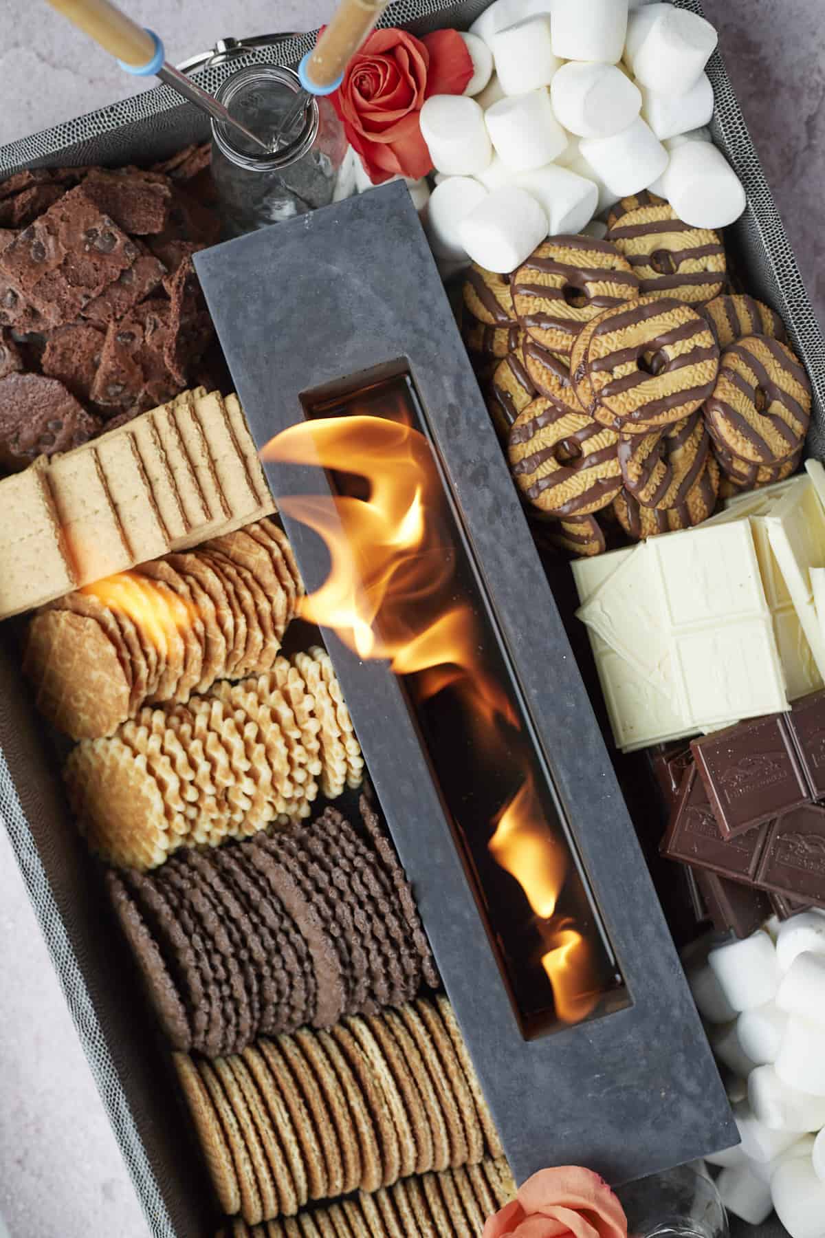 an indoor smores board with a lit fire surrounded by marshmallows, chocolate, cookies, and more