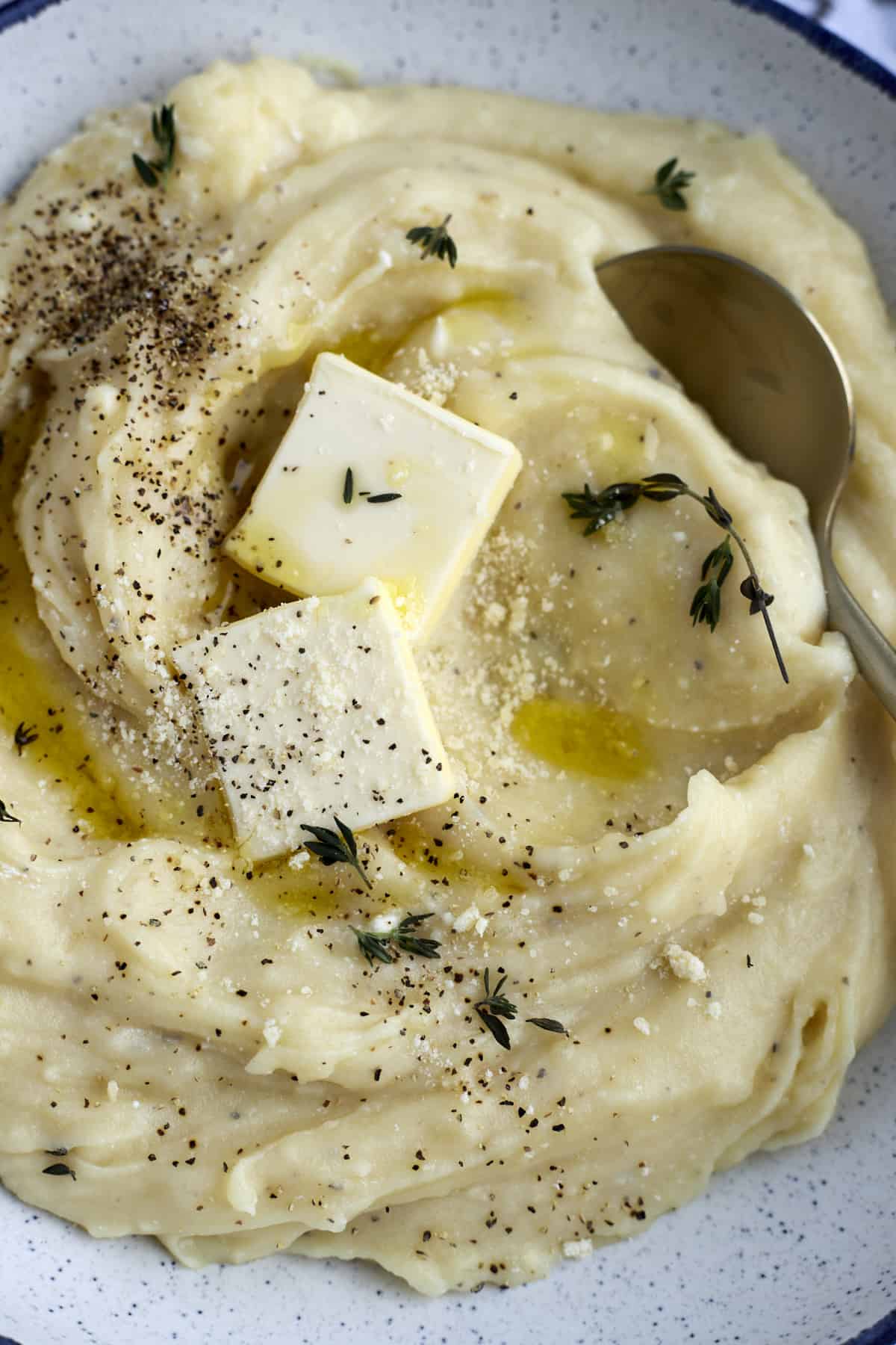 close up image of creamy slow cooker mashed potatoes in a bowl topped with two pads of butter, Parmesan cheese, and pepper