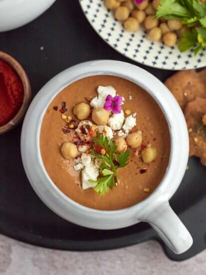 a bowl of spicy tomato soup topped with feta, chickpeas, red pepper flakes, and fresh parsley with more garnishes on the side