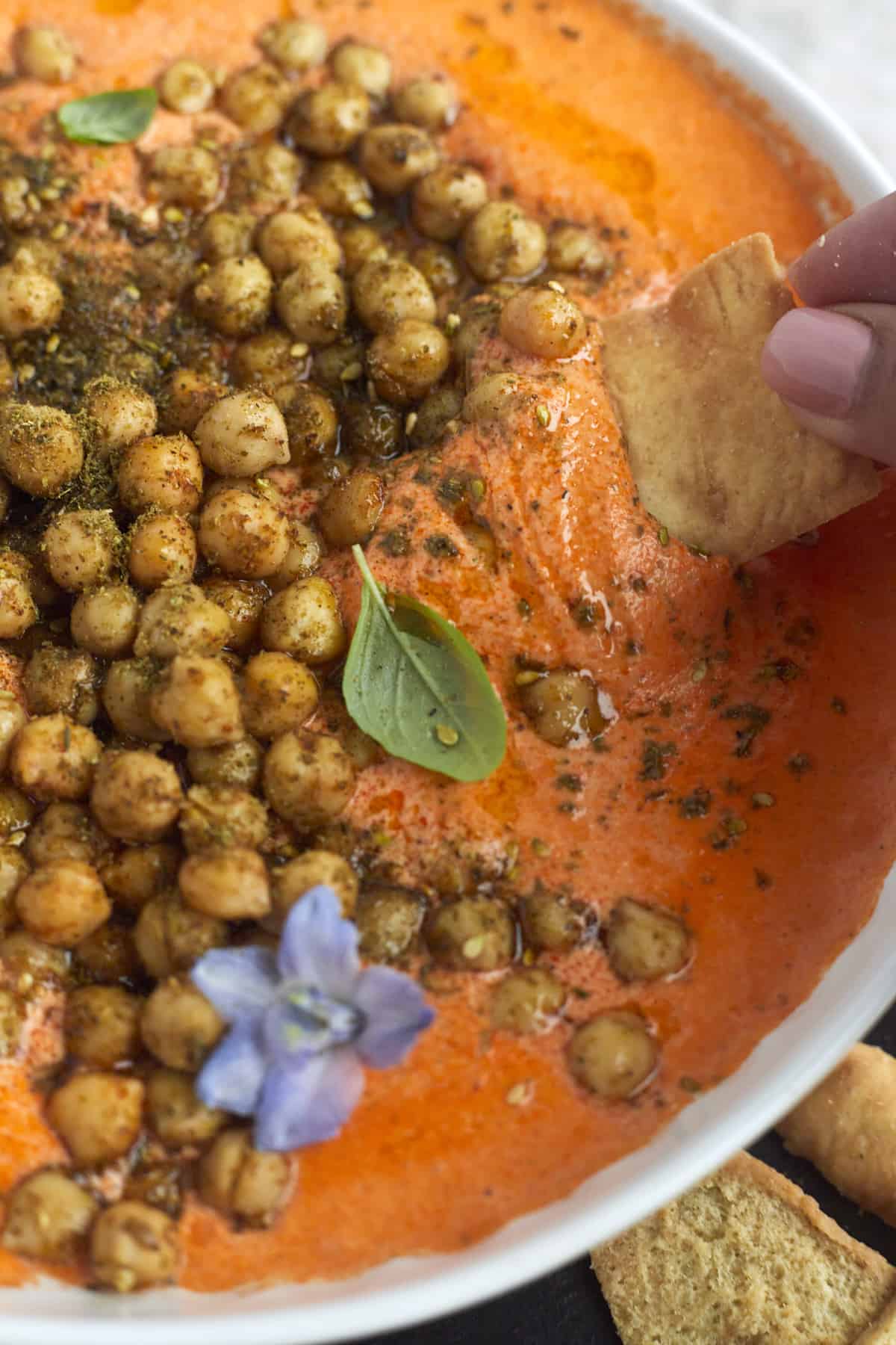 a hand dipping a pita chip into a bowl of Roasted Red Pepper Feta Dip with Crispy Chickpeas