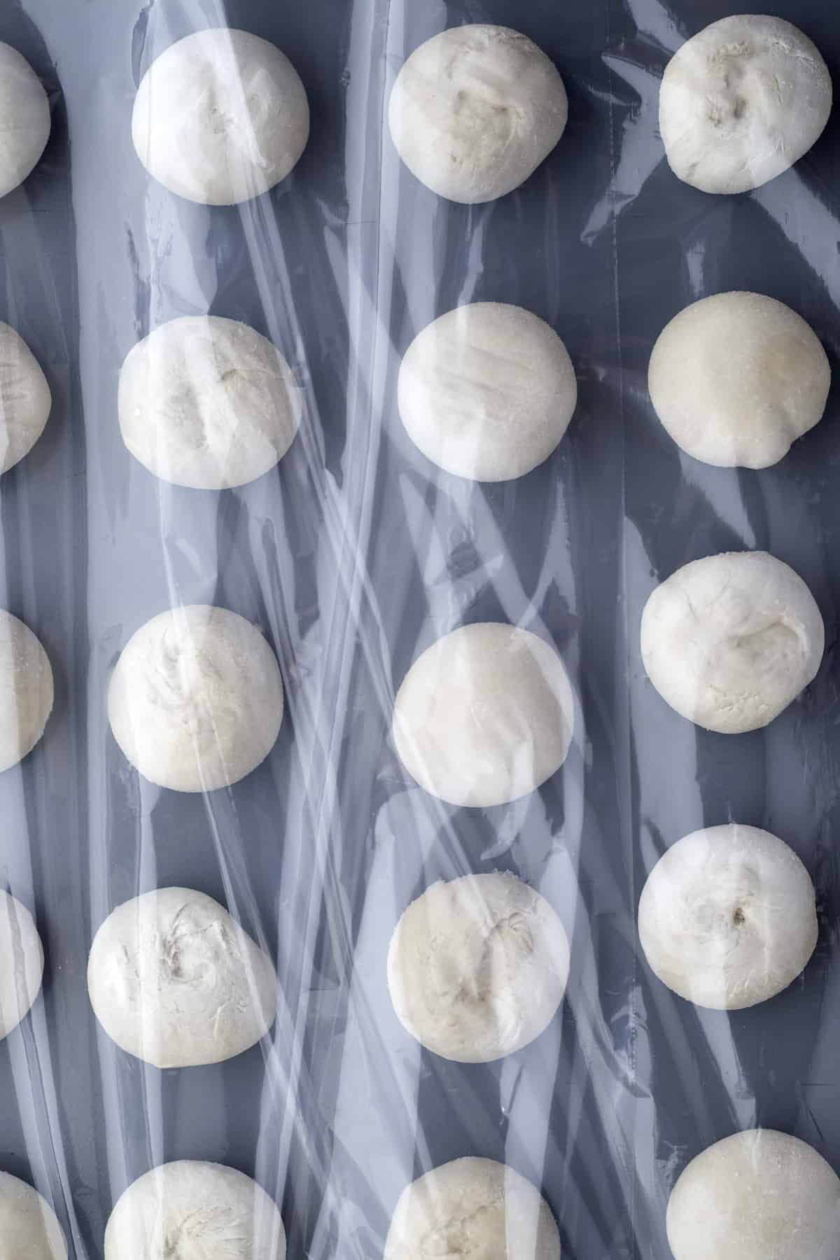 raw bread rolls on a baking sheet covered with plastic wrap