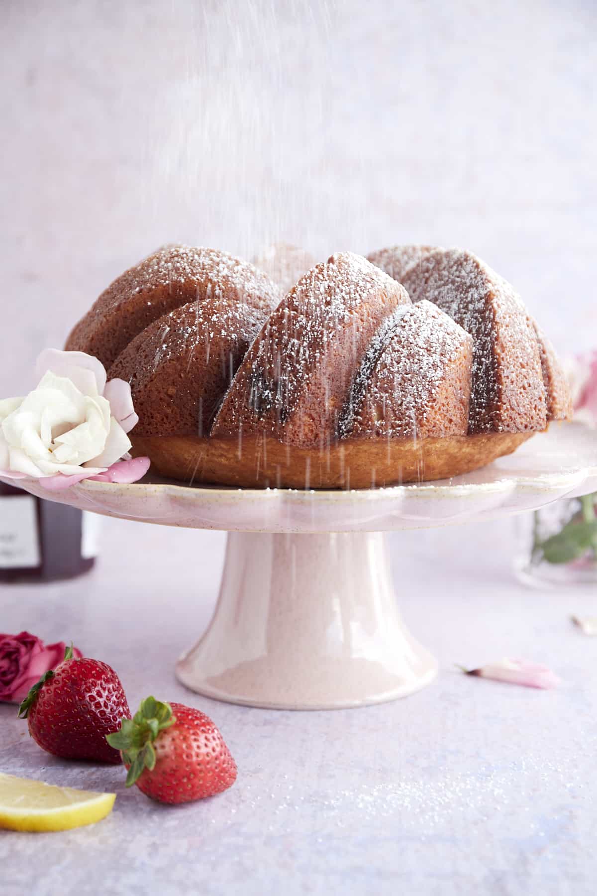 a strawberry lemon bundt cake on a cake stand being dusted with powdered sugar
