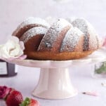 a strawberry lemon bundt cake on a cake stand being dusted with powdered sugar