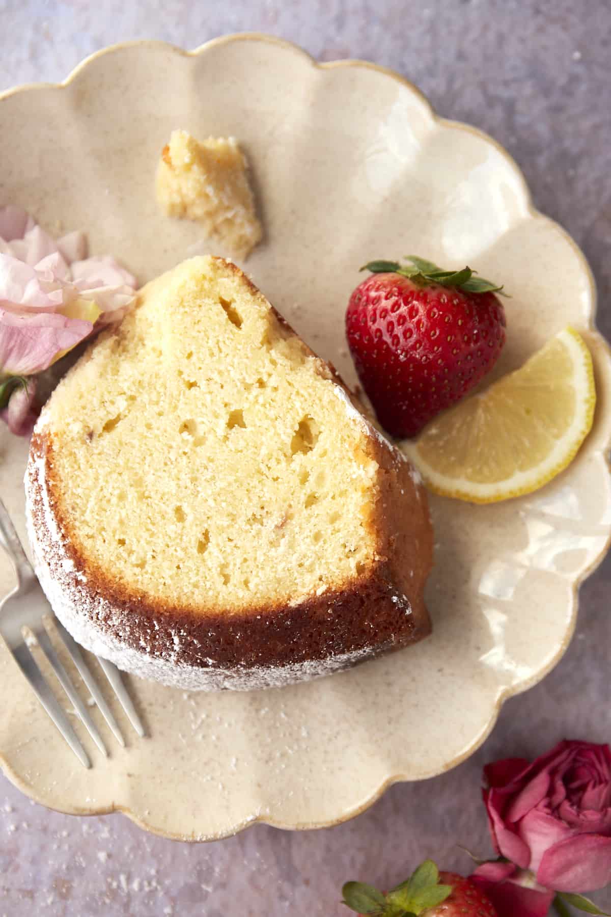 overhead image of a piece of  bundt cake topped with powdered sugar on a plate with a fork, a strawberry, and a lemon slice