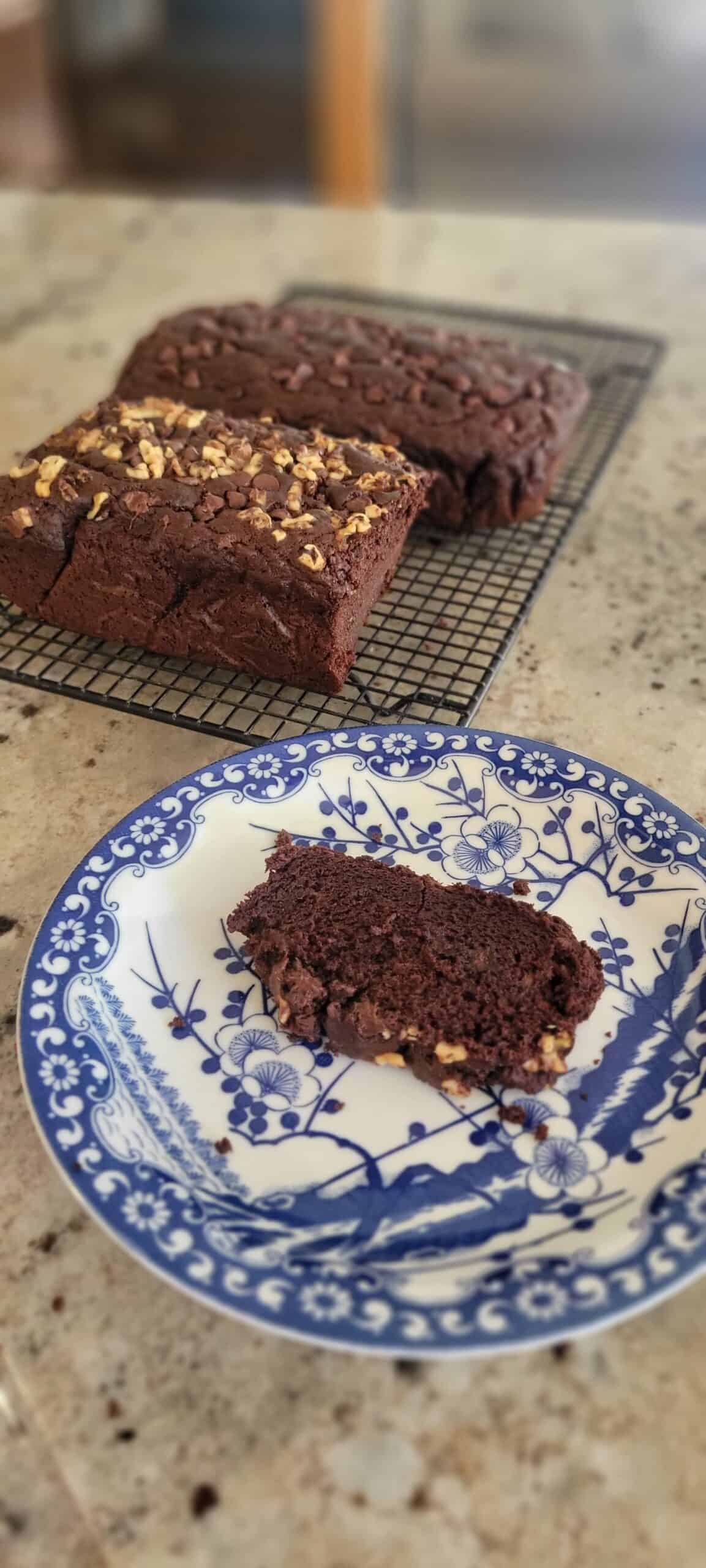 A slice of chocolate loaf cake with walnuts sits on a blue and white floral plate, with two more chocolate loaves on a cooling rack in the background on a kitchen counter.