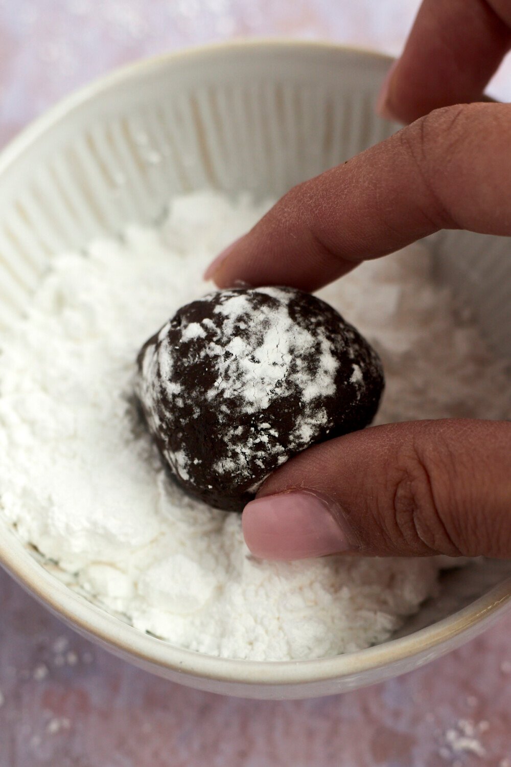 two fingers rolling a ball of chocolate crinkle cookie dough in powdered sugar