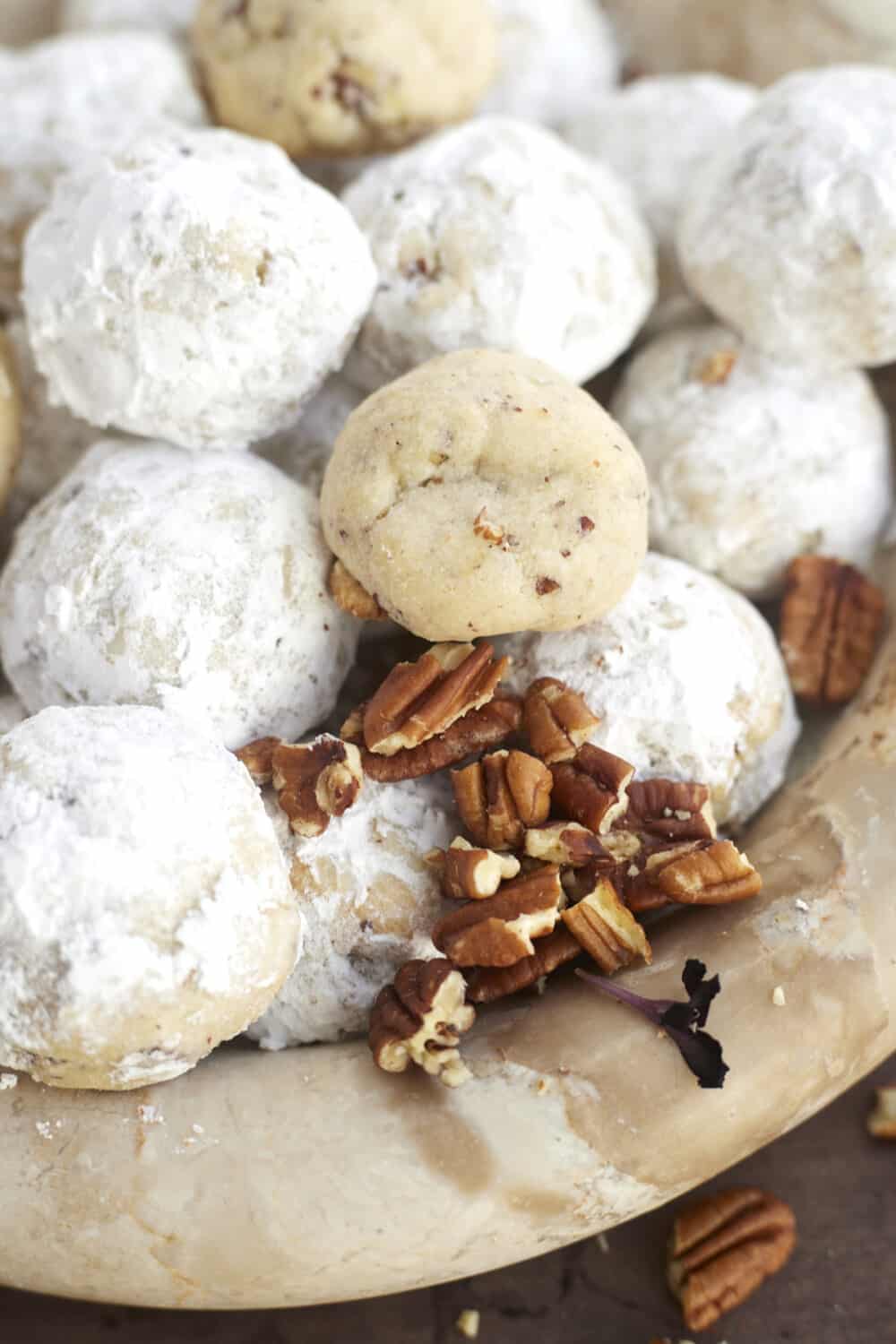 a bowl of snowball cookies with a few not coated in sugar and chopped pecans at the bottom 