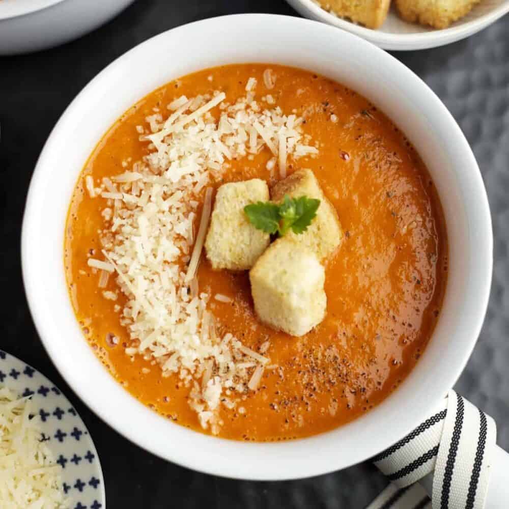 a white bowl full of roasted tomato soup topped with Parmesan cheese and homemade croutons
