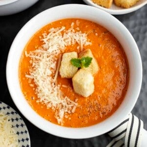 a white bowl full of roasted tomato soup topped with Parmesan cheese and homemade croutons.