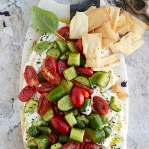 a platter of whipped feta topped with cucumber, roasted tomatoes, and seasonings with pita chips on the side