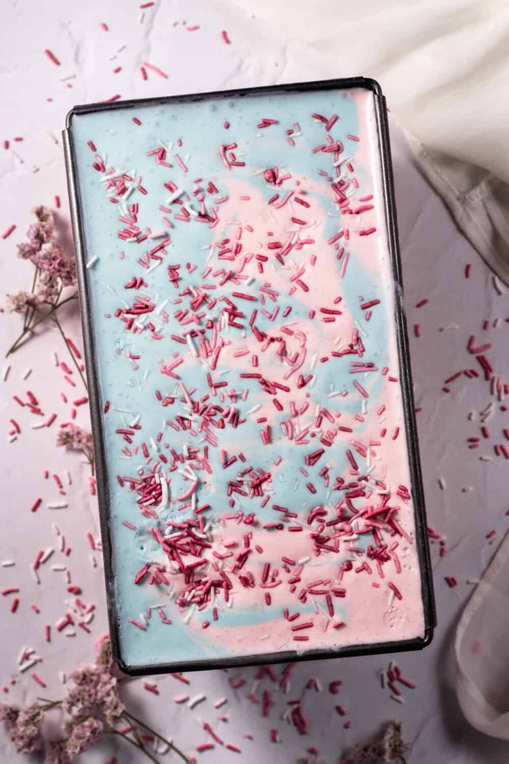 overhead image of a tub of cotton candy ice cream topped with pink and white sprinkles