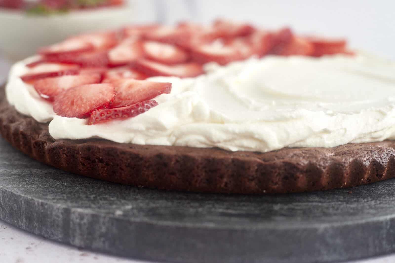 side image of a brownie pizza topped with cream cheese frosting and sliced strawberries