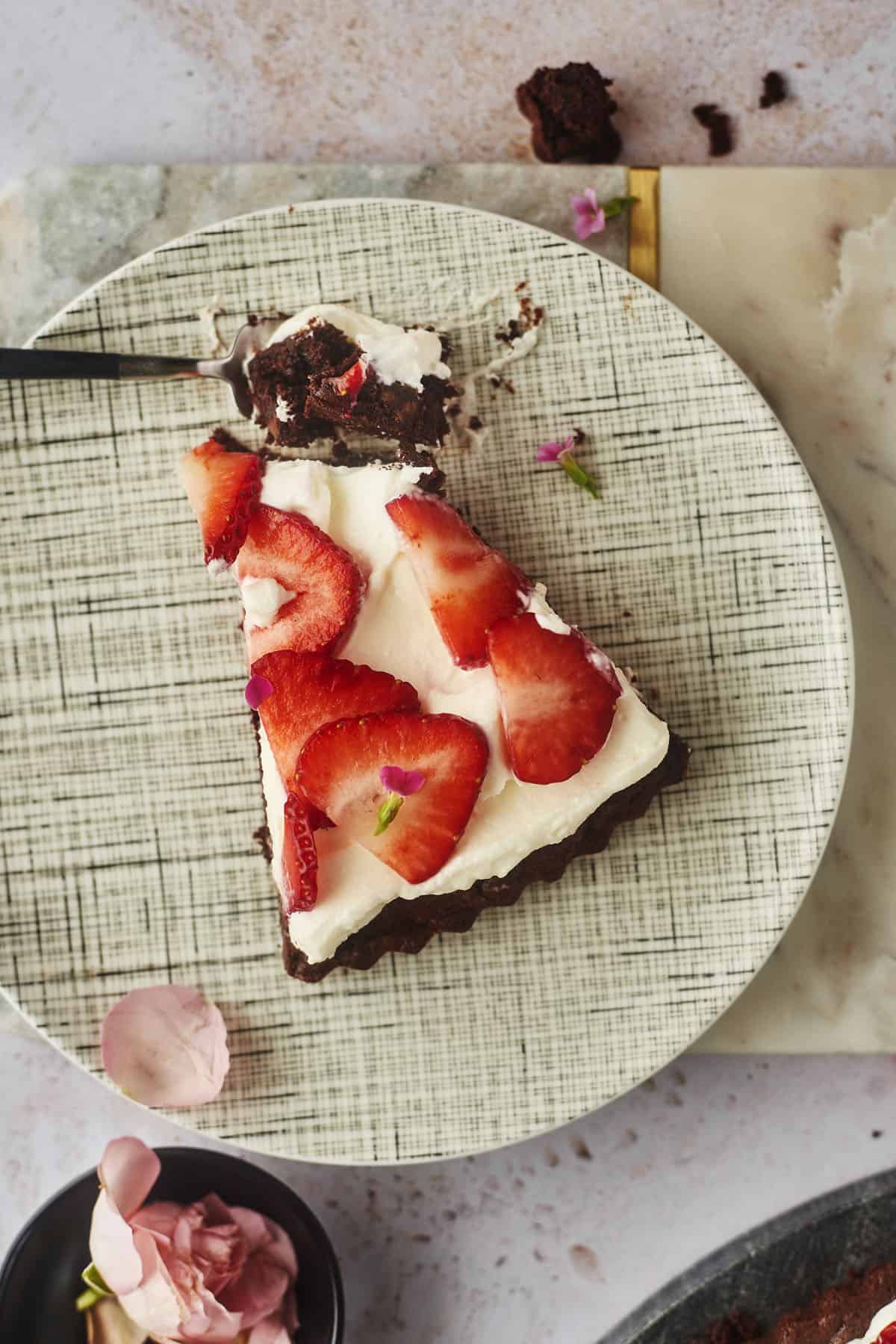 a slice of chocolate brownie pizza topped with cream cheese frosting and strawberries on a plate with a fork full of a bite