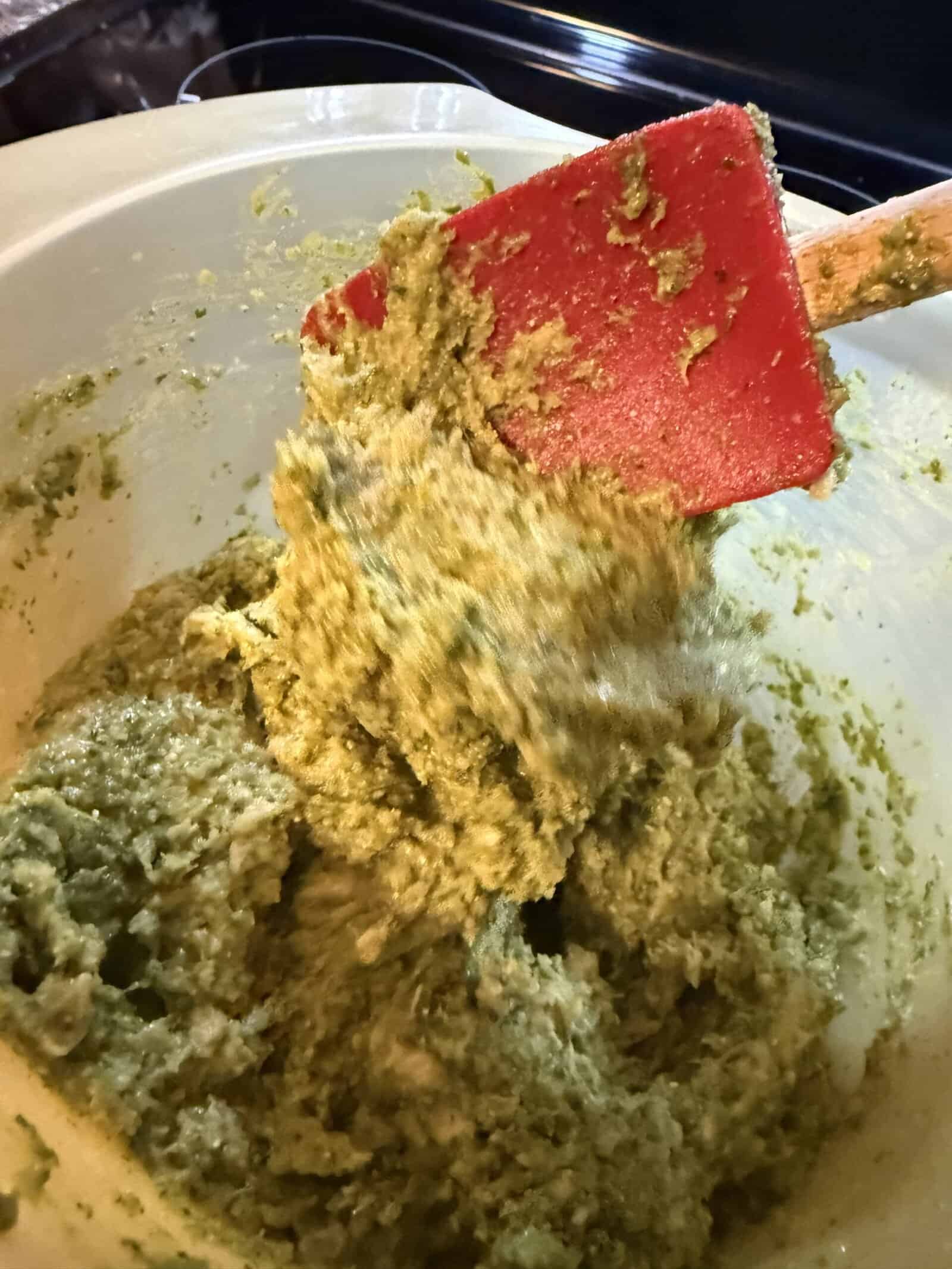 A red spatula stirs a thick, green batter or dough mixture in a large white mixing bowl. The textured mixture appears partially blended.