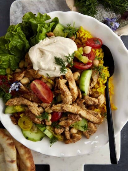 a bowl of shawarma chicken with rice, tomatoes, cucumbers, lettuce, chickpeas, and hummus
