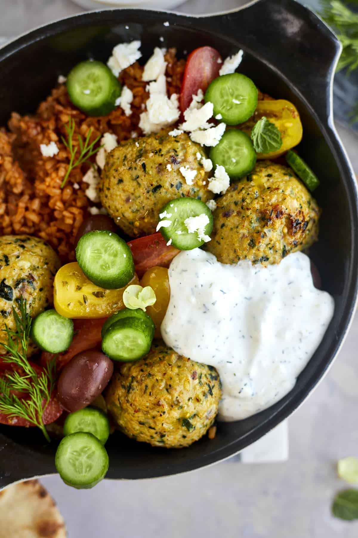 Greek meatballs in a skillet with rice, cucumbers, tomatoes, tzatziki, and feta
