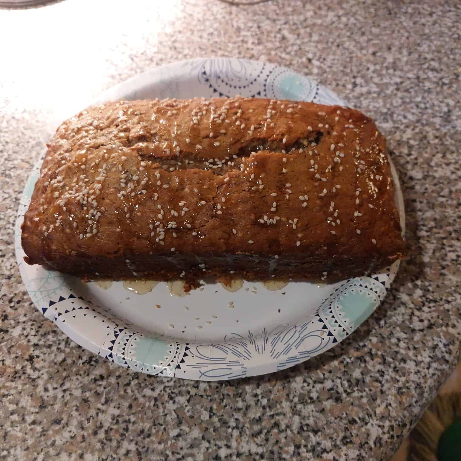 A loaf of homemade bread topped with sesame seeds sits on a patterned paper plate, placed on a speckled kitchen countertop.