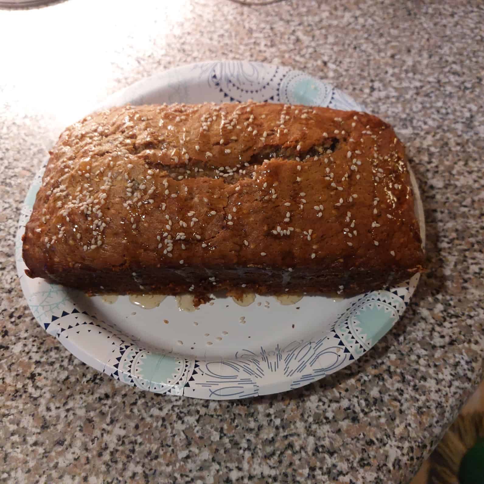 A loaf of bread topped with oats sits on a decorative paper plate on a speckled countertop. The bread has a golden-brown crust and a crack running along the top.