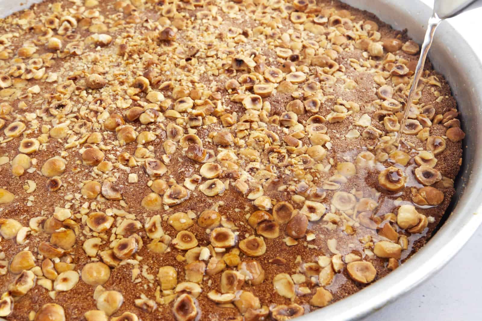 Simple syrup being poured over baked basbousa topped with hazelnuts. 