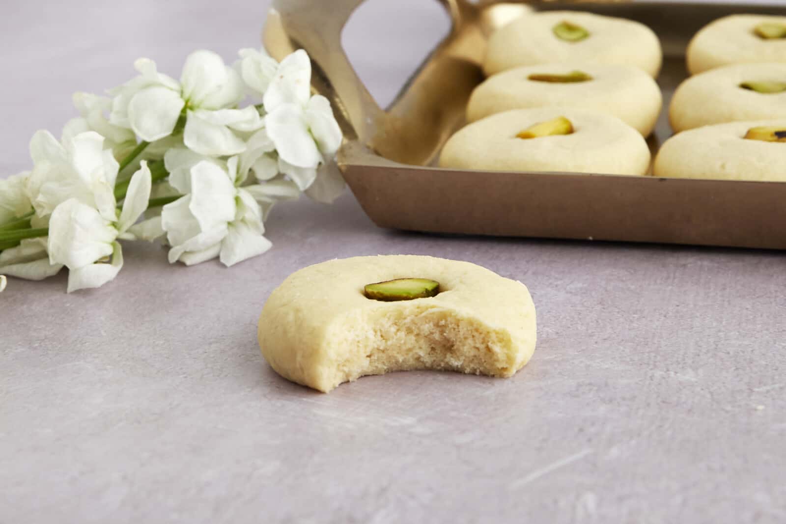 A box of ghraybeh cookies topped with pistachios with one on the counter missing a bite. 