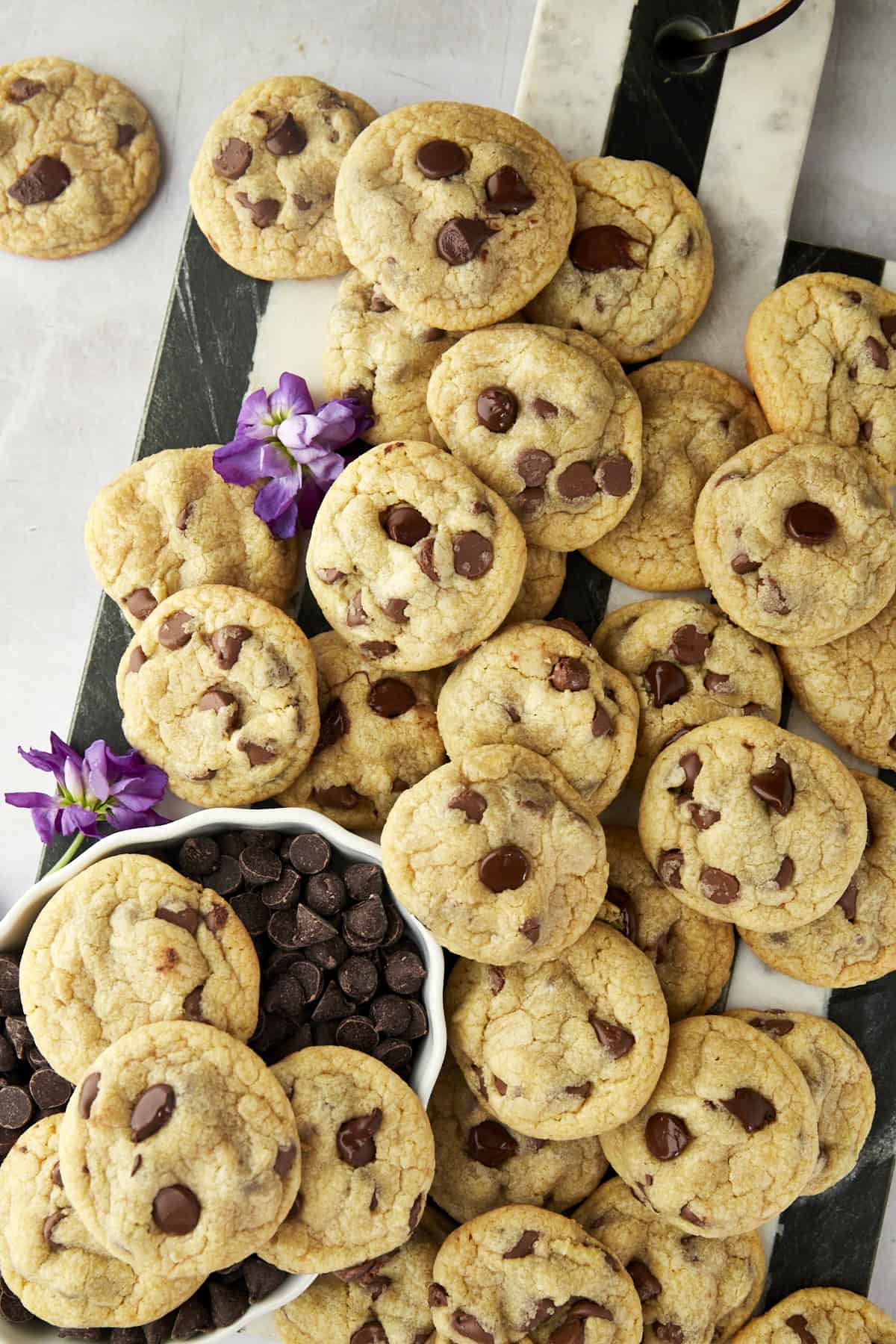 A pile of chocolate chip cookies is spread on a marble and wood board with purple flowers and a small bowl of chocolate chips beside them.