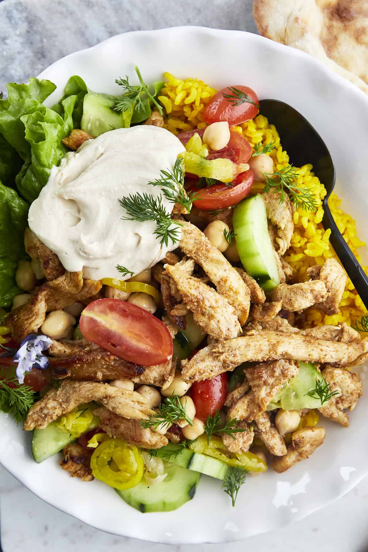 a bowl of shawarma chicken with yellow rice, cucumbers, tomatoes, chickpeas, lettuce, and hummus