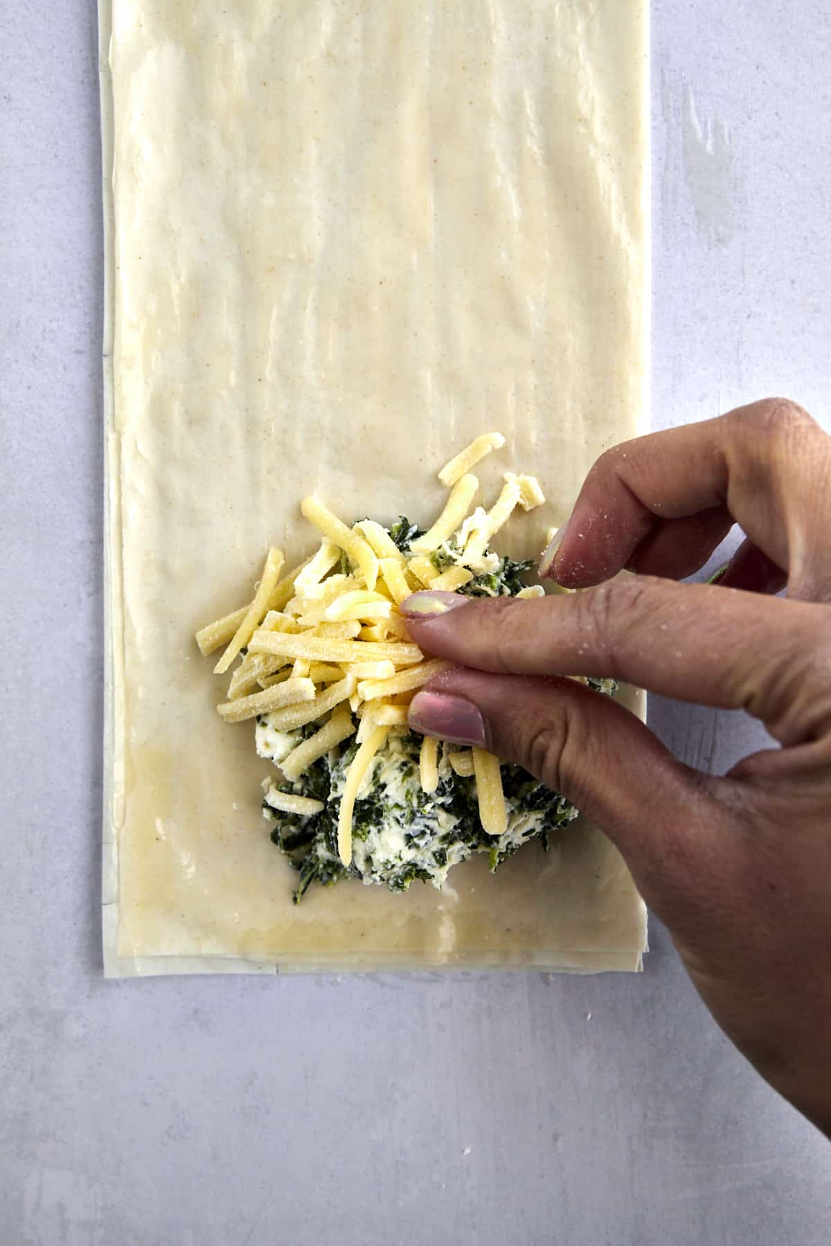 A hand sprinkling cheese over a spinach spanakopita filling over layered phyllo dough. 