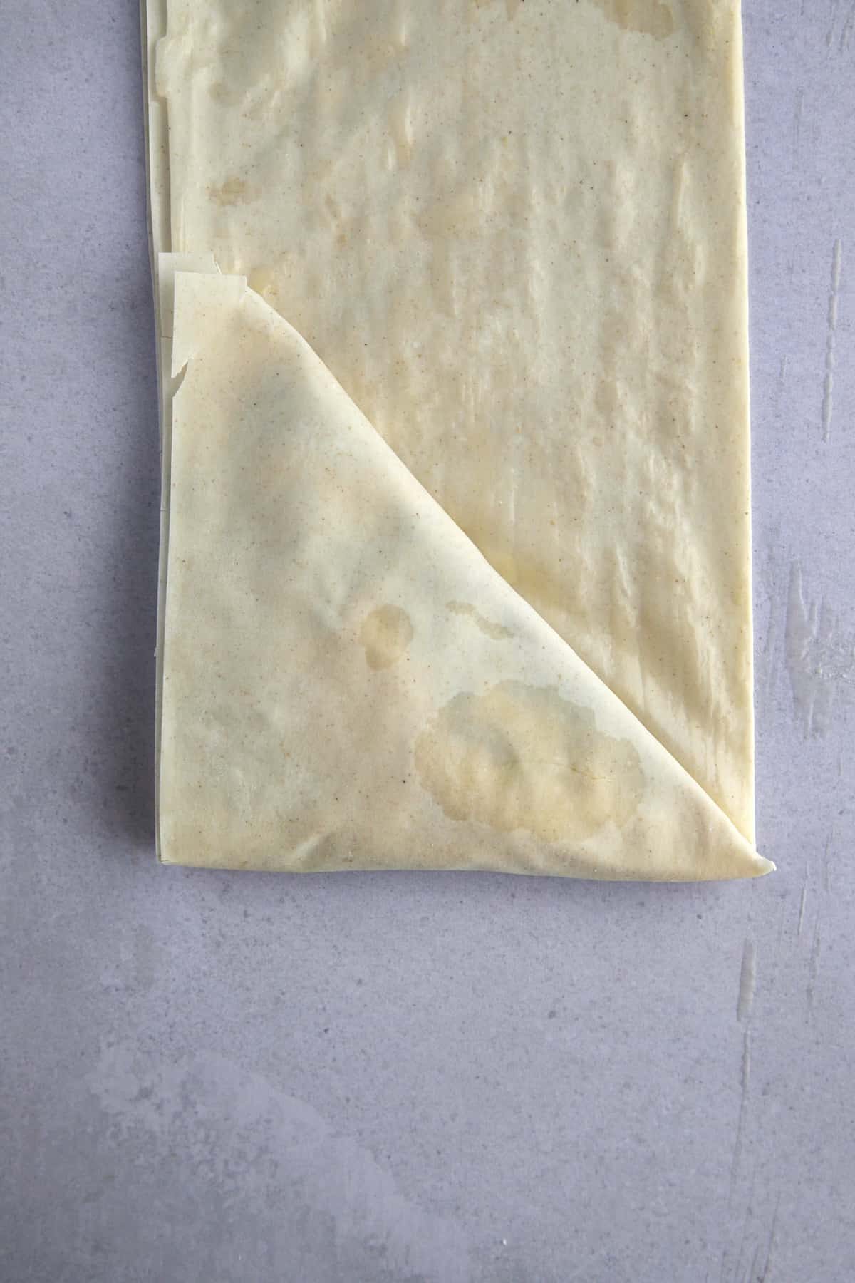 Raw layered phyllo dough sheets stuffed with spanakopita filling being folded into a triangle. 