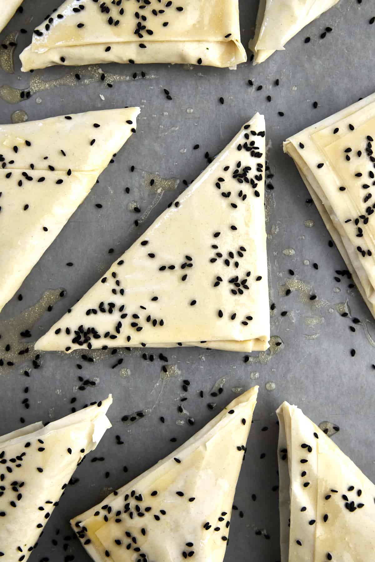 Raw spanakopita triangles on a baking sheet topped with butter and black sesame seeds. 