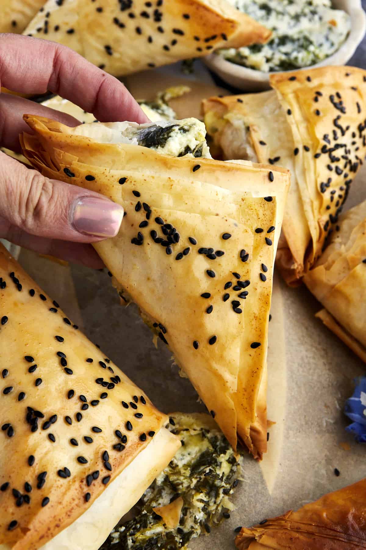 A hand holding a baked spanakopita triangle topped with sesame seeds. 