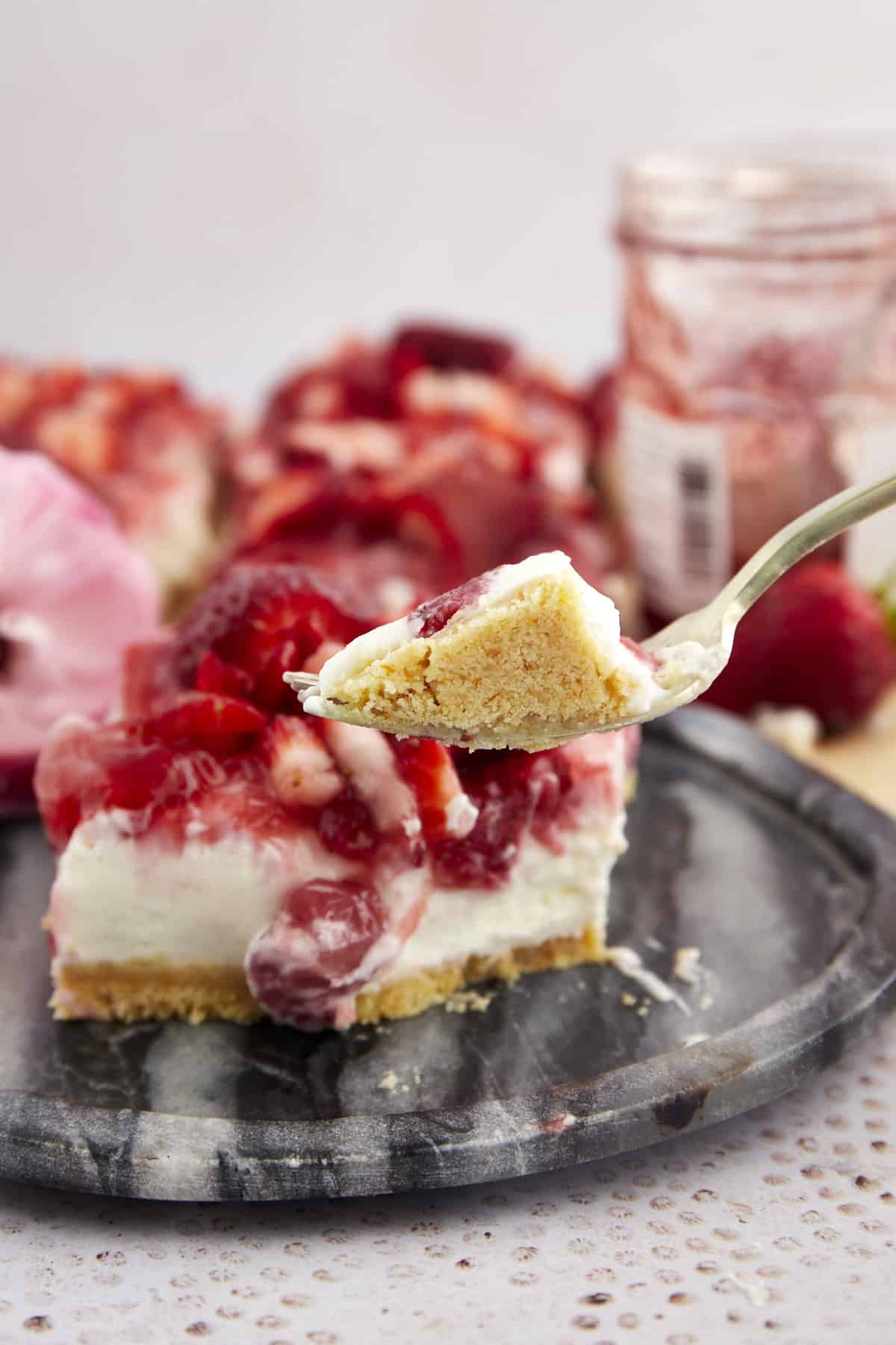 A fork holding a bite of a strawberry cheesecake bar. 