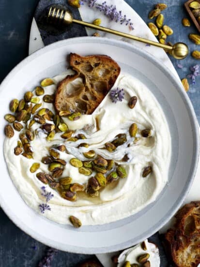 A bowl of whipped ricotta topped with roasted pistachios and honey with a piece of crostini.