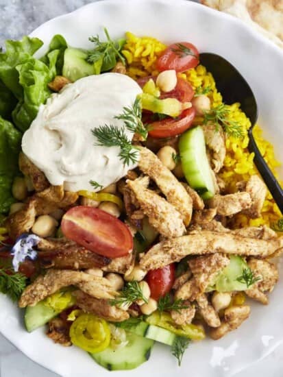 a bowl of shawarma chicken with yellow rice, tomatoes, cucumber, chickpeas, lettuce, and hummus