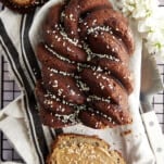 A loaf of tahini banana bread topped with honey and sesame seeds with a slice cut off and topped with nut butter and extra sesame seeds.