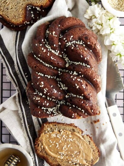 A loaf of tahini banana bread topped with honey and sesame seeds with a slice cut off and topped with nut butter and extra sesame seeds.