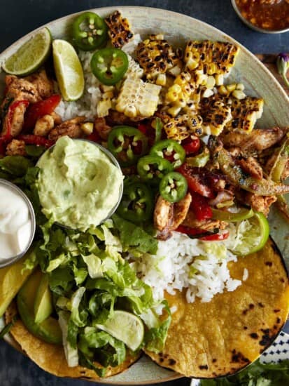 A chicken fajita bowl with rice, grilled corn, mashed avocado, sour cream, lettuce, and corn tortillas.