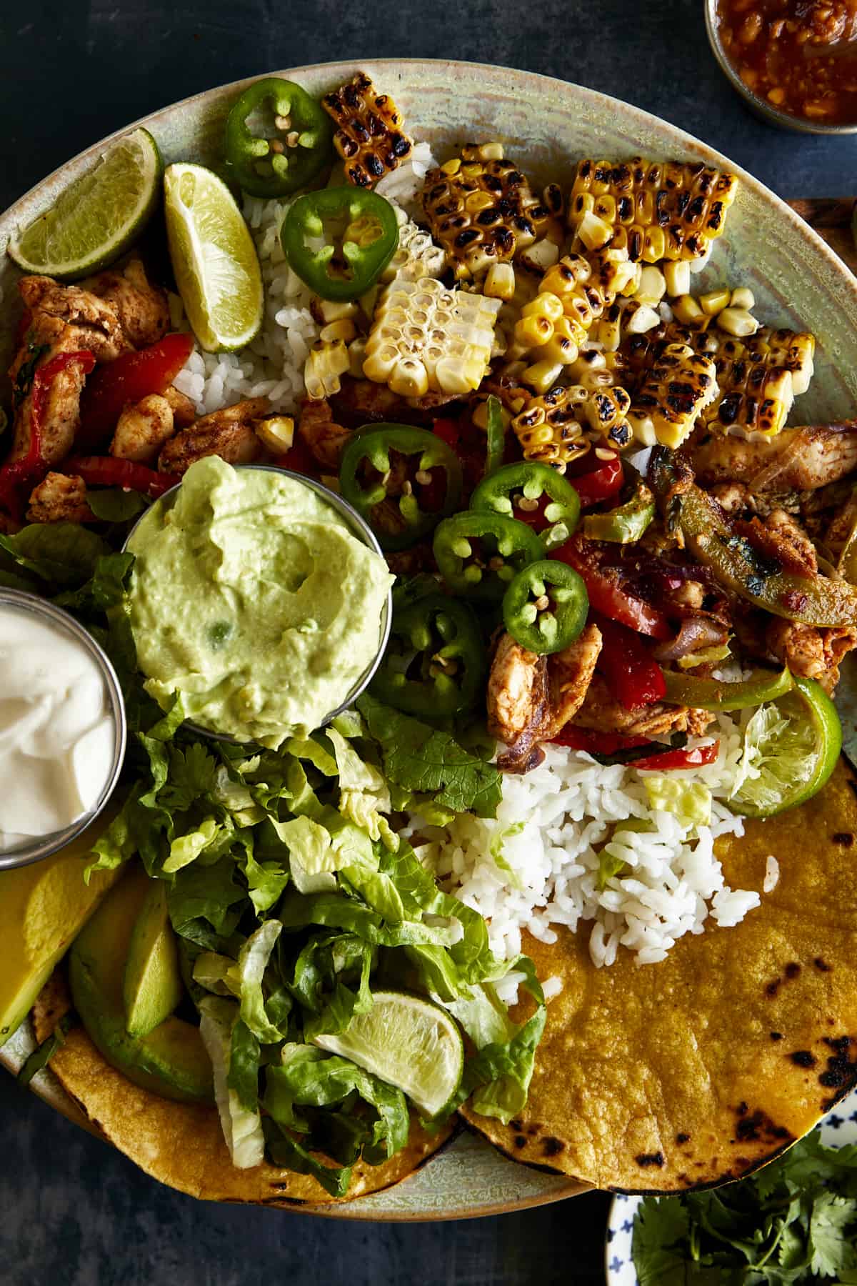 A chicken fajita bowl with chicken, peppers and onions, grilled corn, cilantro lime rice, mashed avocado, sour cream, corn tortillas, and lettuce. 