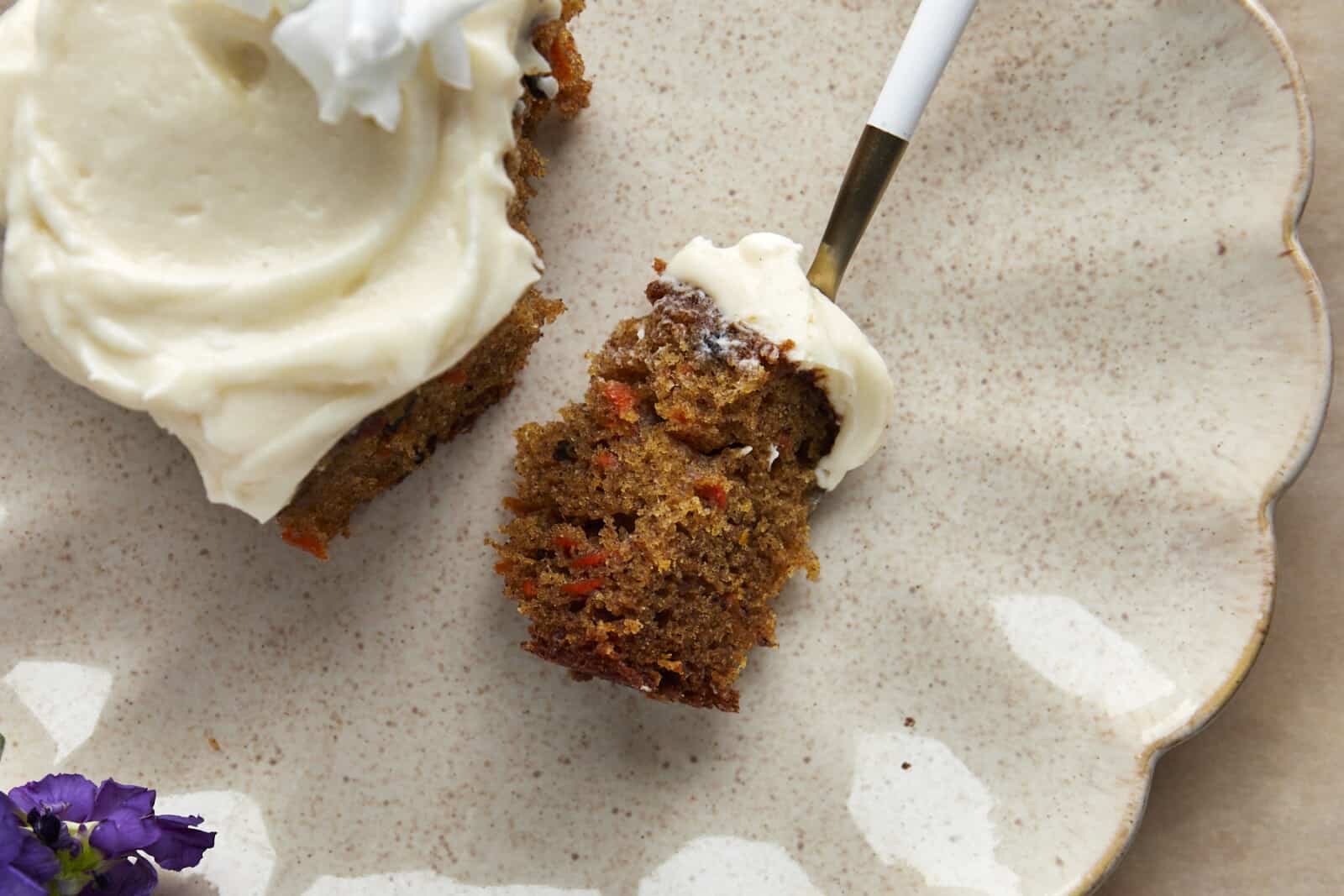 A forkful of carrot cake topped with vanilla bean cream cheese frosting on a plate with the rest of the piece of carrot cake. 