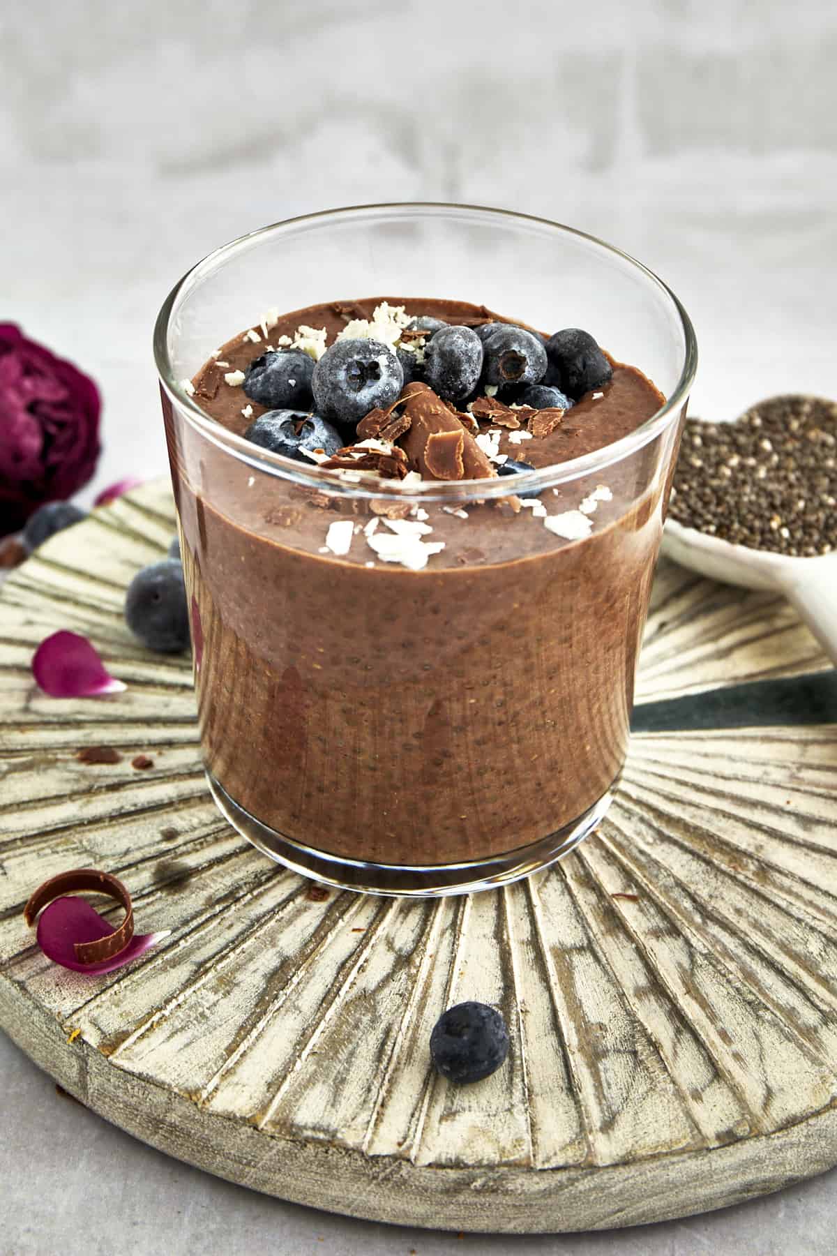 A jar of chocolate chia pudding topped with blueberries, chocolate shavings, and coconut flakes with a spoonful of chia seeds in the background. 