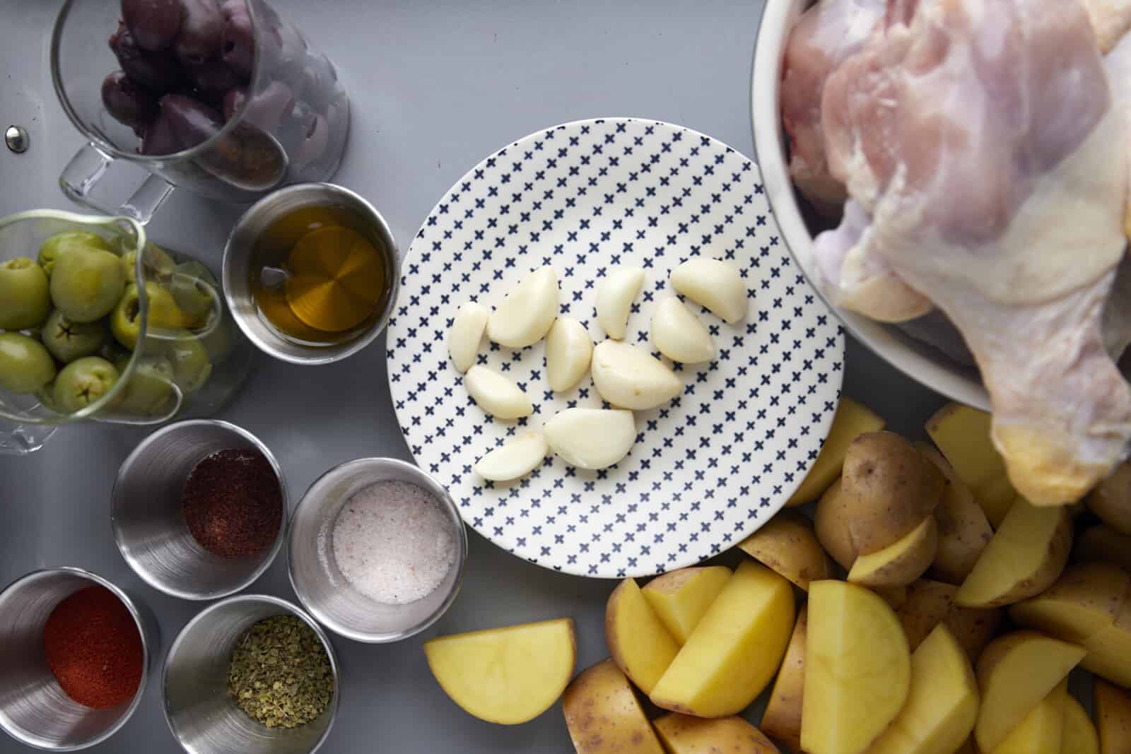 Raw chicken drumsticks, golden potatoes, garlic cloves, spices, olive oil, green olives, and kalamata olives. 