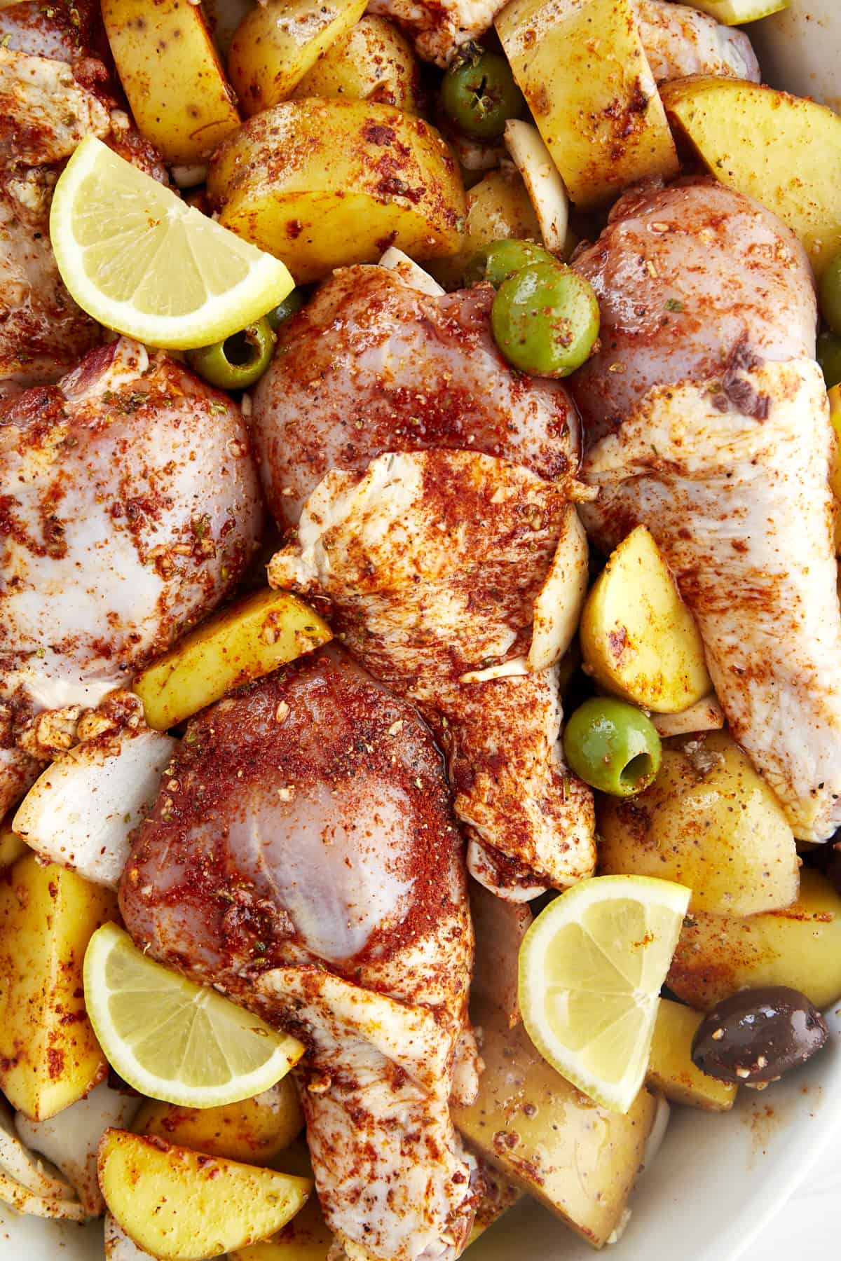Raw chicken drumsticks with golden potatoes, red and green olives, onions, and garlic coated with seasonings and topped with lemon wedges. 