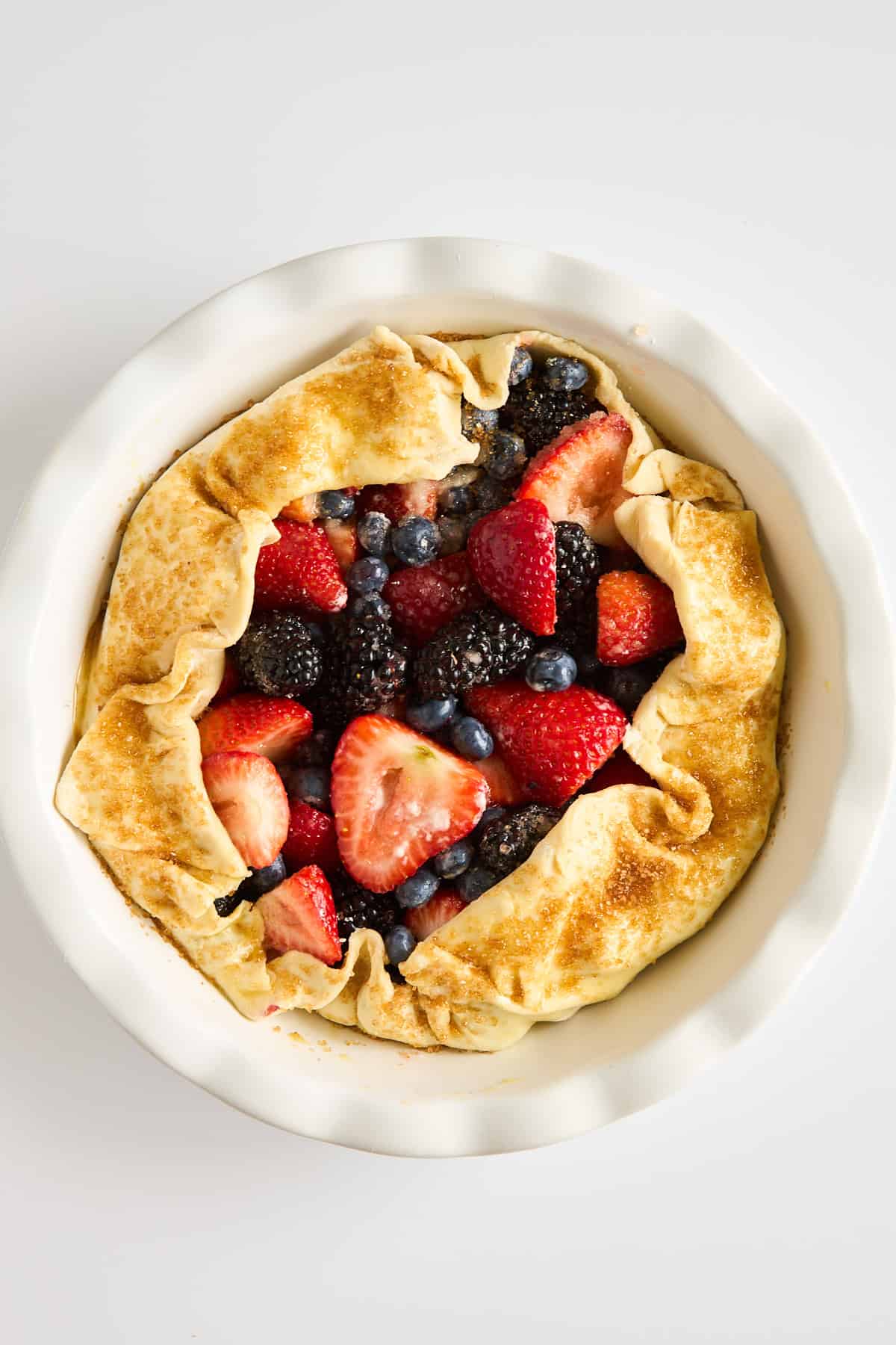 A rustic galette with a golden, folded crust filled with fresh strawberries, blueberries, and blackberries, sits in a white pie dish on a white background.