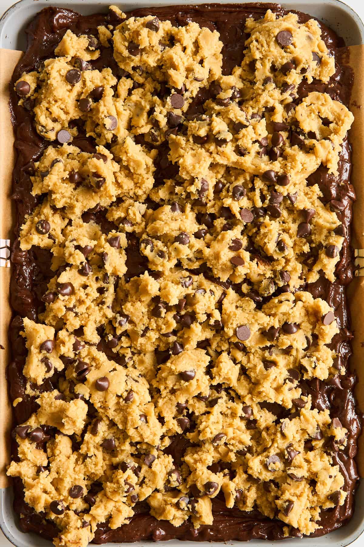 A baking tray filled with brownie batter topped with uneven scoops of chocolate chip cookie dough, ready to be baked.
