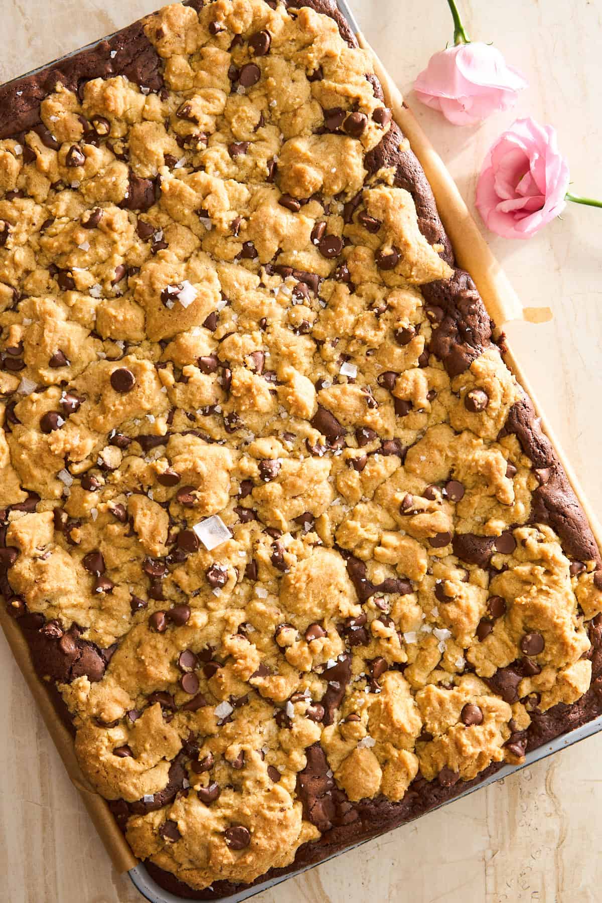 A large tray of chocolate chip cookie bars with a golden, crumbly topping and chocolate chips, set on parchment paper with two pink flowers nearby on a light surface.