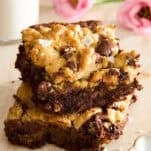 Two chocolate chip cookie bars stacked on a light surface, sprinkled with flaky sea salt. A glass of milk and pink flowers are in the blurred background.