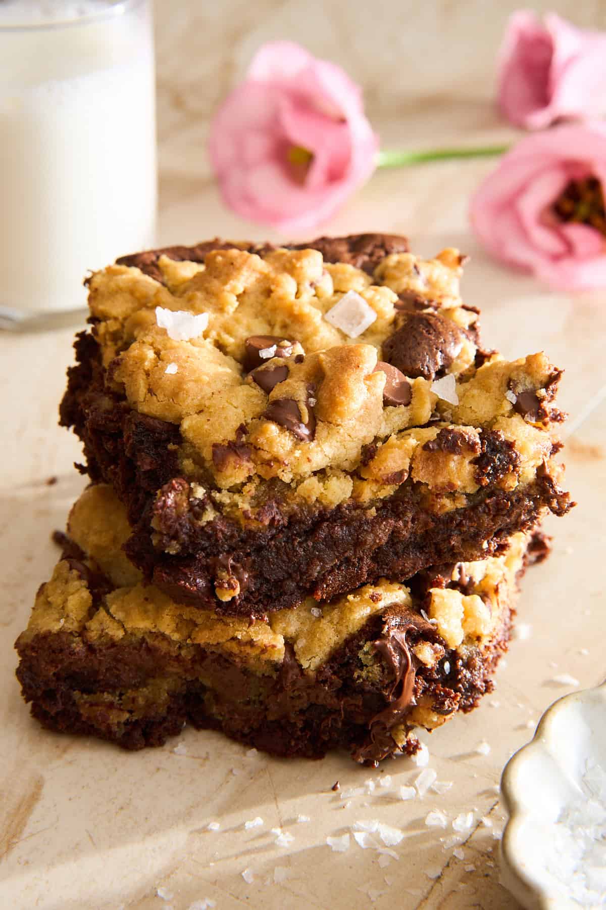 Two chocolate chip cookie bars stacked on a light surface, sprinkled with flaky sea salt. A glass of milk and pink flowers are in the blurred background.